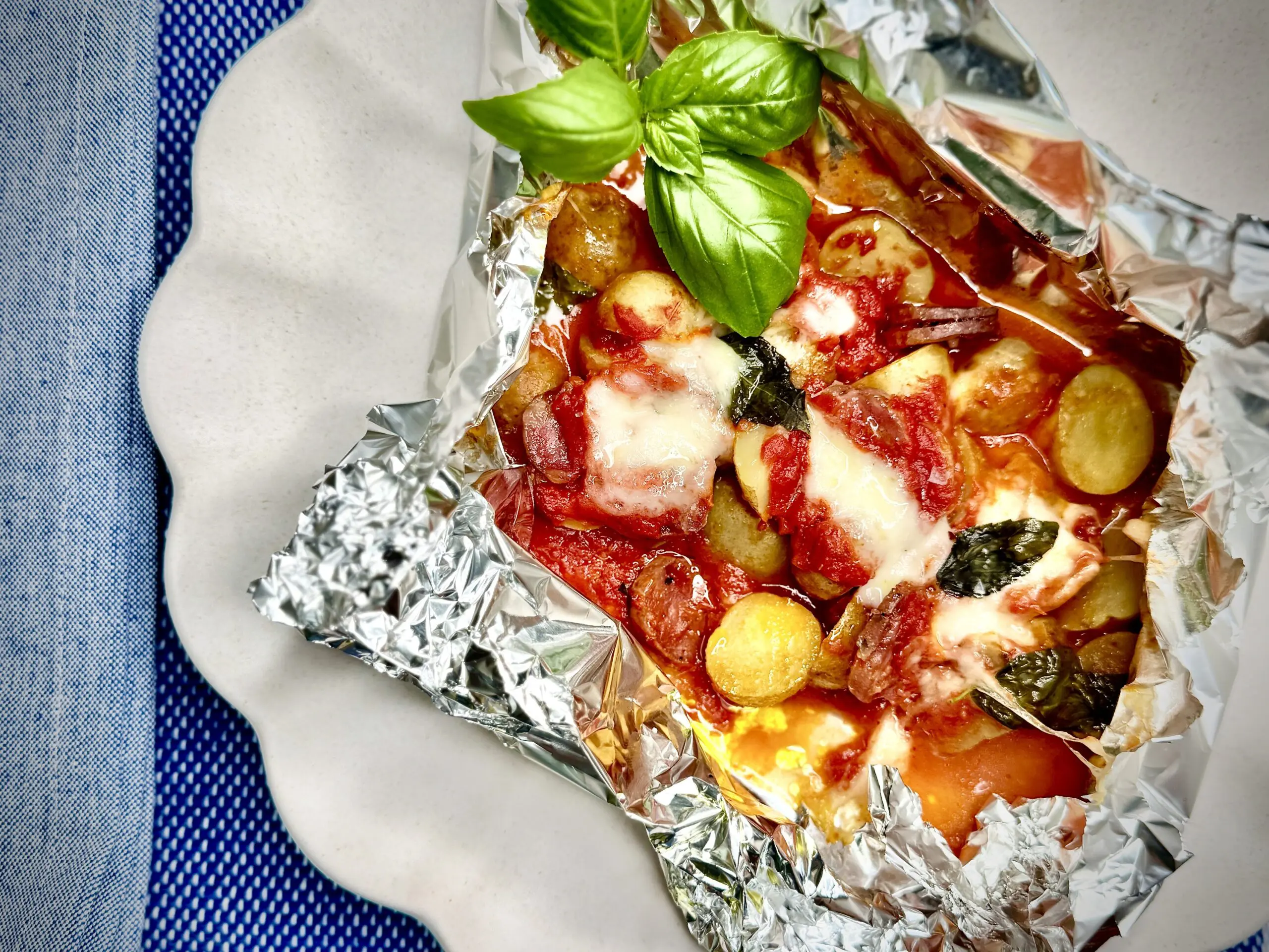 A white plate with aluminum foil on it and inside the aluminum foil is such a delicious assortment of pizza sauce, Little Potatoes, and shredded melty cheese.