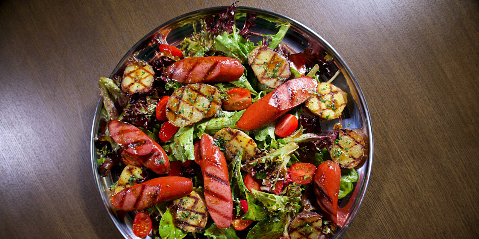 A delicious salad featuring Little Potatoes and grilled sausage sits on a plate against a wooden table.
