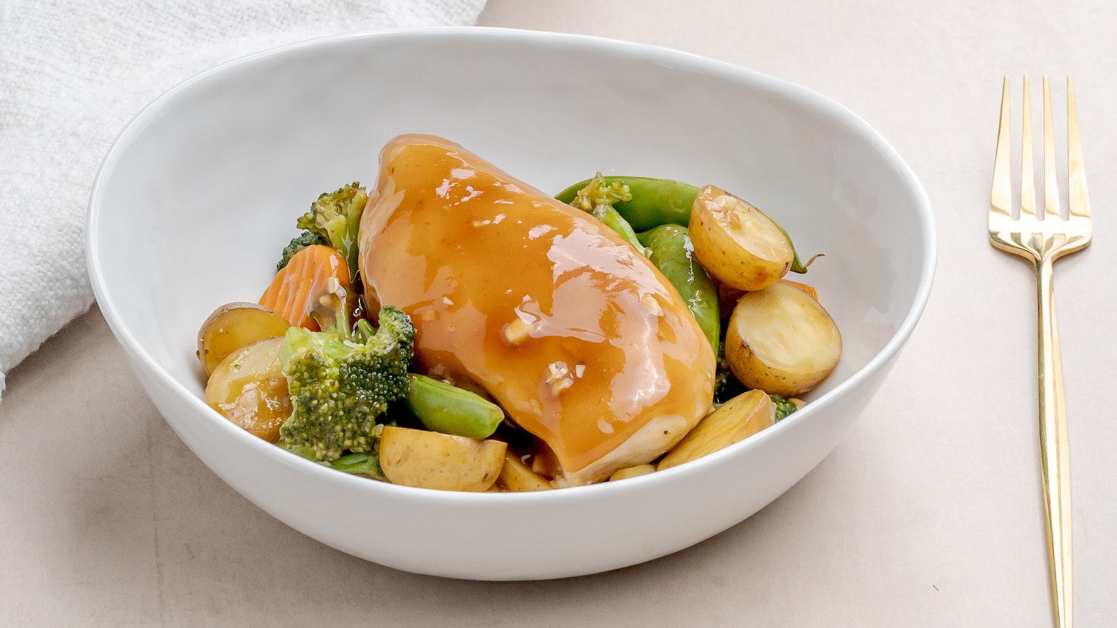 Tender chunks of chicken, Little Potatoes, and colorful vegetables coated in a honey garlic sauce, served in a white bowl with fresh herbs on top.