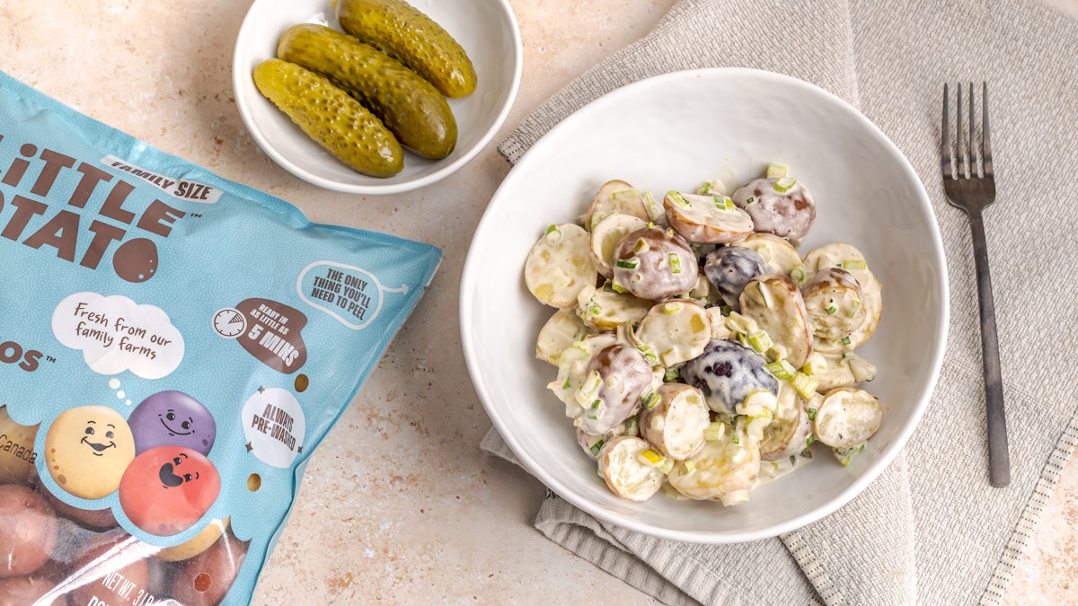 Dill pickle potato salad in a white bowl with another bowl of pickles nearby on a table.