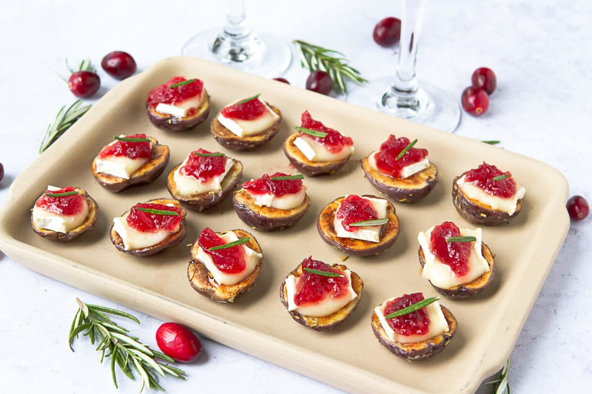 Roasted Little Potato bites topped with melted brie and cranberry sauce, arranged on a platter and garnished with fresh herbs.
