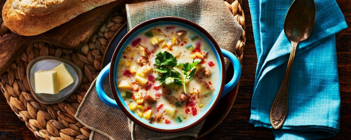 A bowl of potato and corn chowder with some butter and crusty bread nearby.