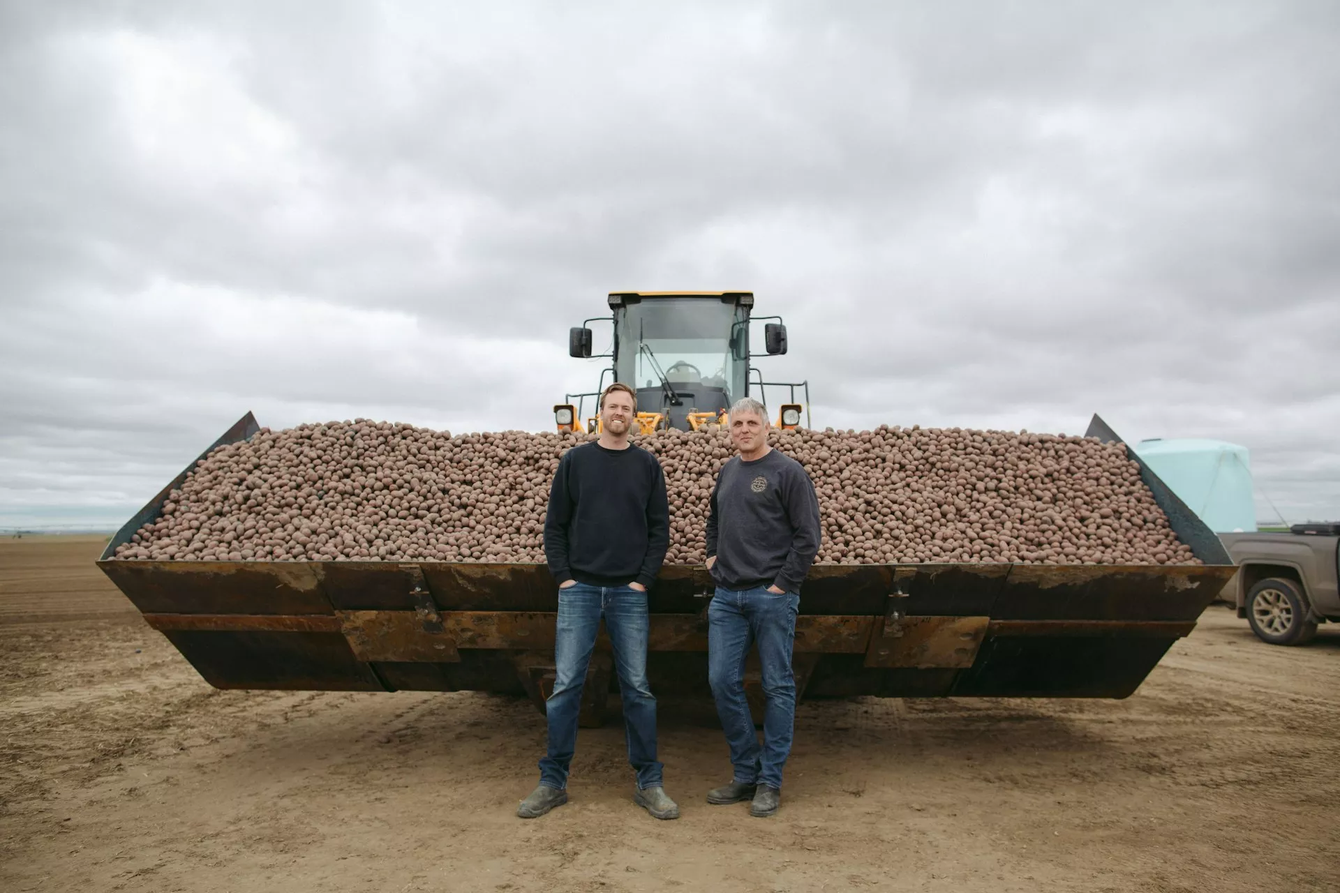 Two farmers stand in front of a pile of potatoes they've grown.