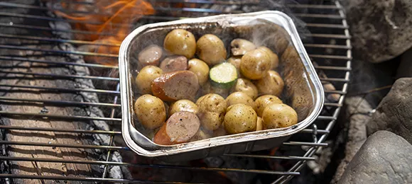 A foil pan full of little potatoes and veggies over a campfire.