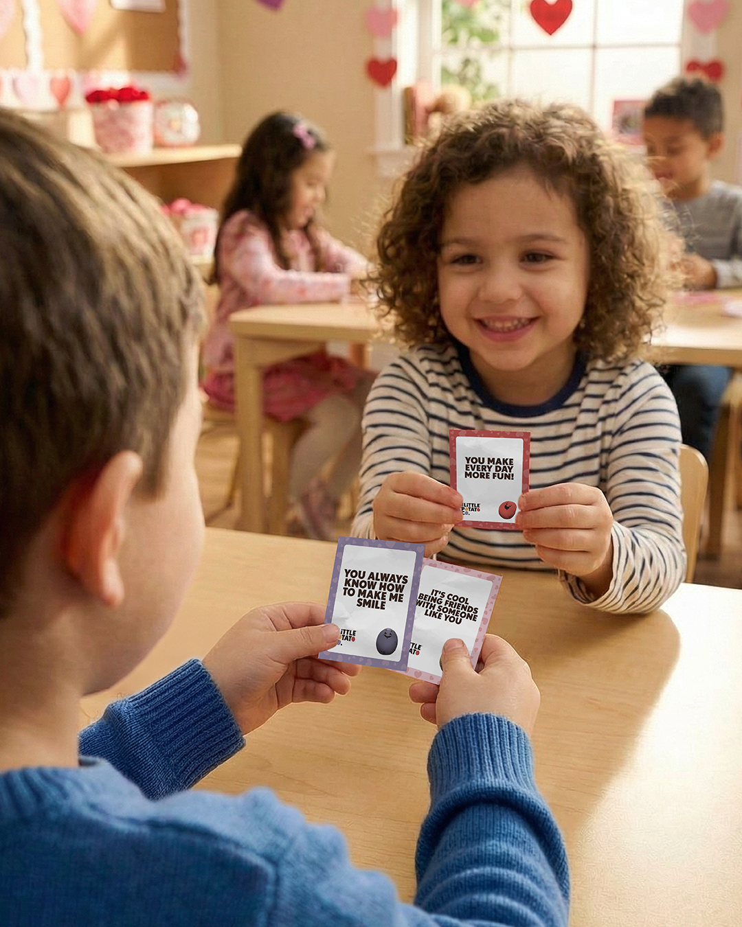 Children sit around a table at school holding Little Potato Company themed Valentines' that say things like 'You always know how to make me smile.'