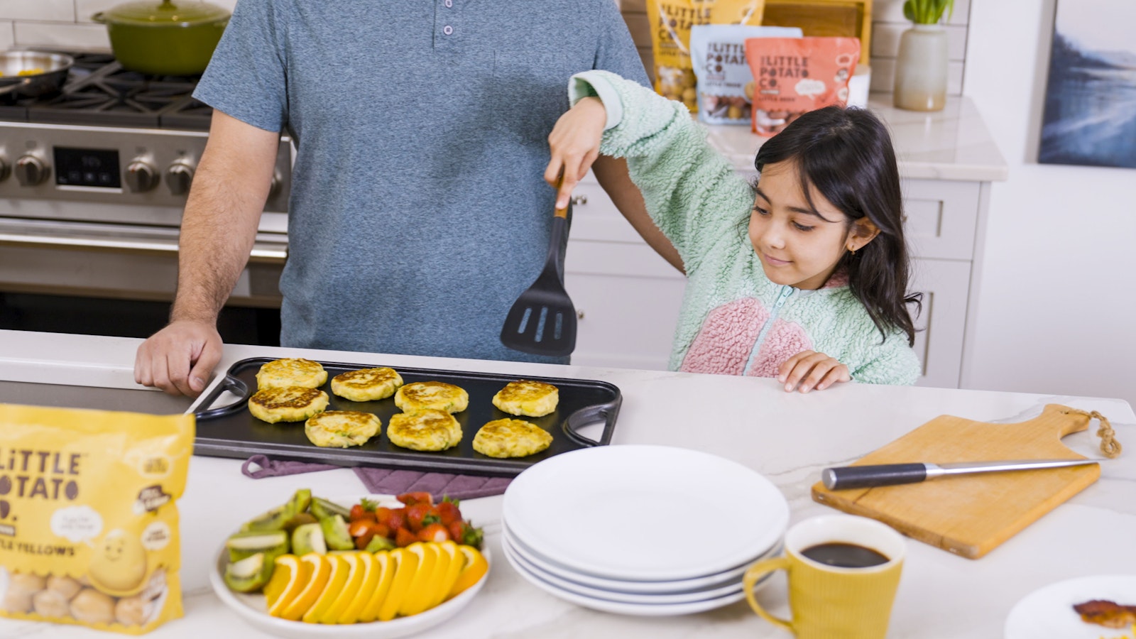 A child stands next to her parent in the kitchen, using a spatula to scoop up a breakfast patty from a baking sheet.